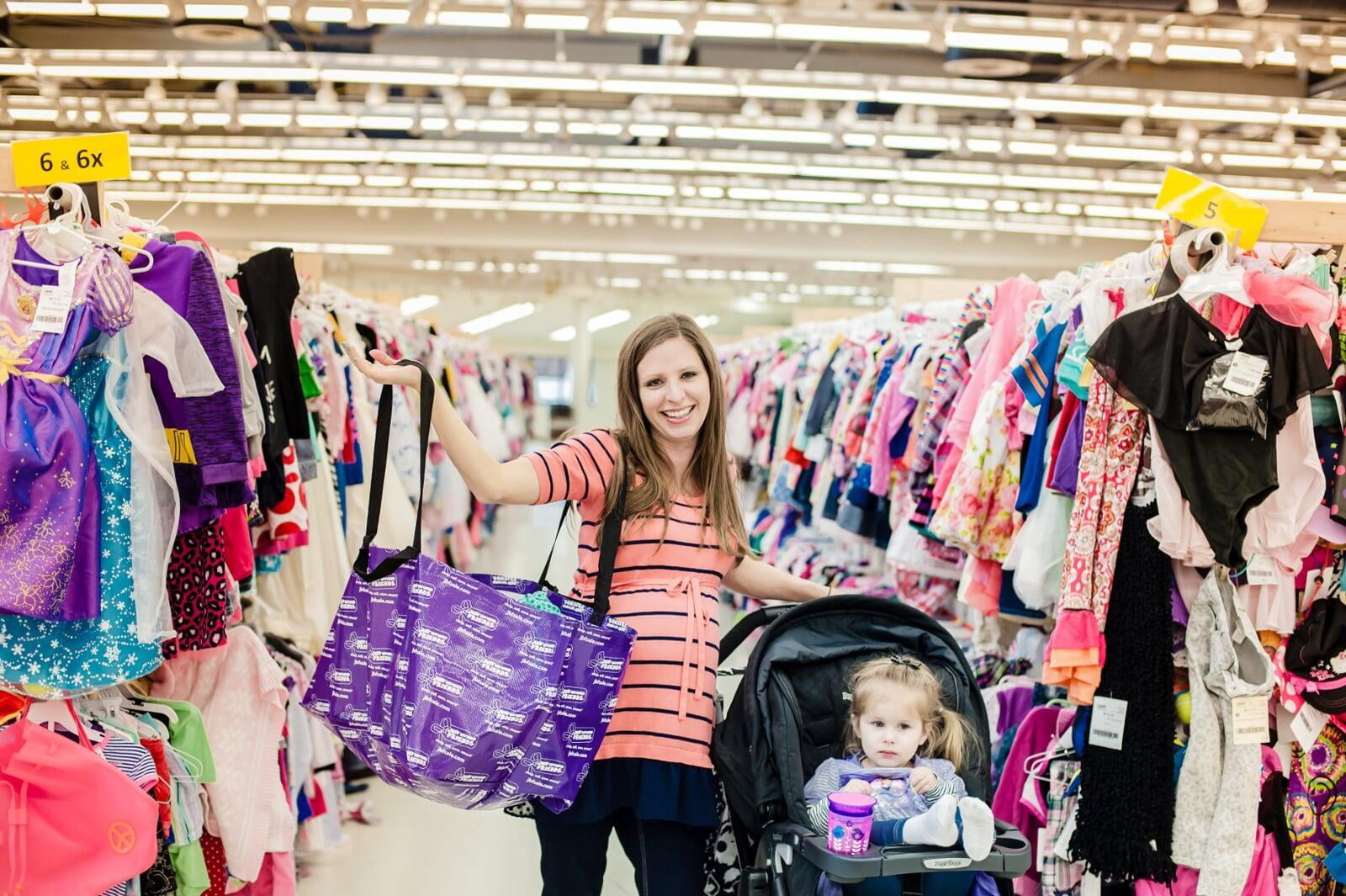 A Mom holds up her JBF shopping bag with her right hand while resting her left hand on her stroller with toddler daughter inside.